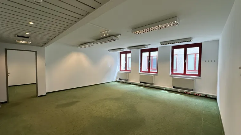 Geräumiger Büroraum mit grünem Teppichboden, weißen Wänden und mehreren Fenstern für Tageslicht.