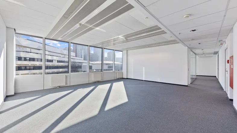 Geräumiges, leeres Büro mit großen Fenstern, grauem Teppichboden und viel Tageslicht.