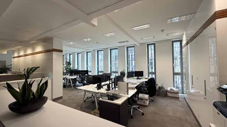 Geräumiges Großraumbüro mit mehreren Arbeitsplätzen, großen Fenstern und Teppichboden.