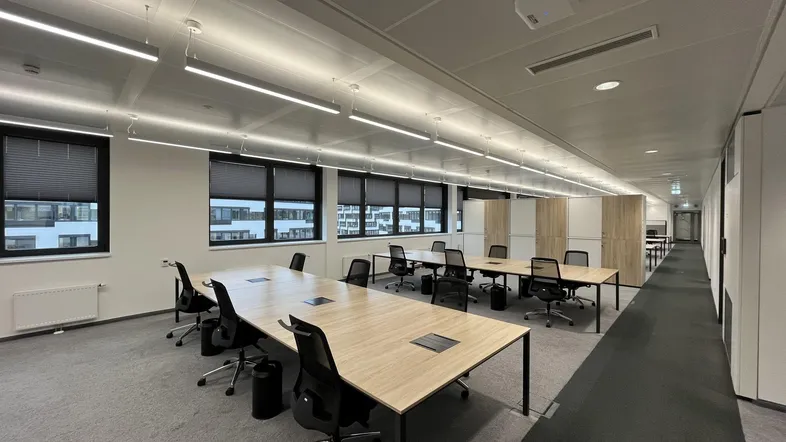 Moderner Großraumbüro-Arbeitsbereich mit langen Holztischen, ergonomischen Stühlen und großen Fenstern mit Jalousien.