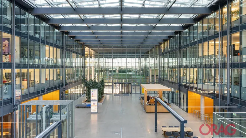 Großzügiges, modernes Atrium eines Bürogebäudes mit Glasdach, mehreren Etagen und einem Empfangsbereich.