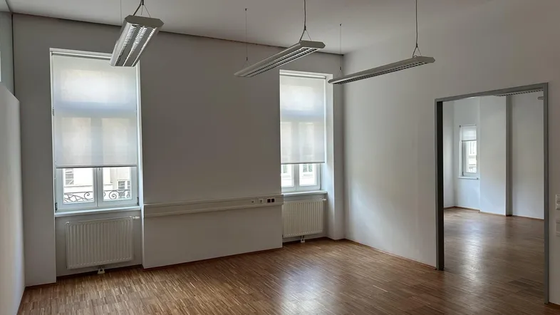 Heller Büroraum mit Holzparkettboden, weißen Wänden und großen Fenstern mit Rollos.