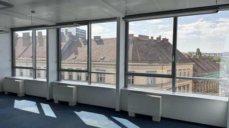 Geräumiges Büro mit großen Fenstern, blauem Teppichboden und Blick auf die Dächer der Stadt.