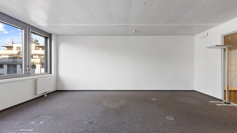 Leerer Büroraum mit Teppichboden, großen Fenstern und Blick auf umliegende Gebäude.