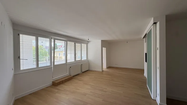 Helles Wohnzimmer mit Parkettboden, weißen Wänden und großen Fenstern mit Jalousien.