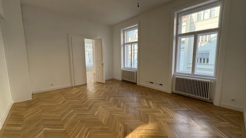 Geräumiges Zimmer mit Fischgrätparkett, weißen Wänden und großen Fenstern, die viel Tageslicht hereinlassen.