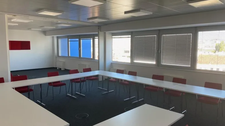 Konferenzraum mit U-förmiger Tischordnung, roten Stühlen und großen Fenstern mit Jalousien.