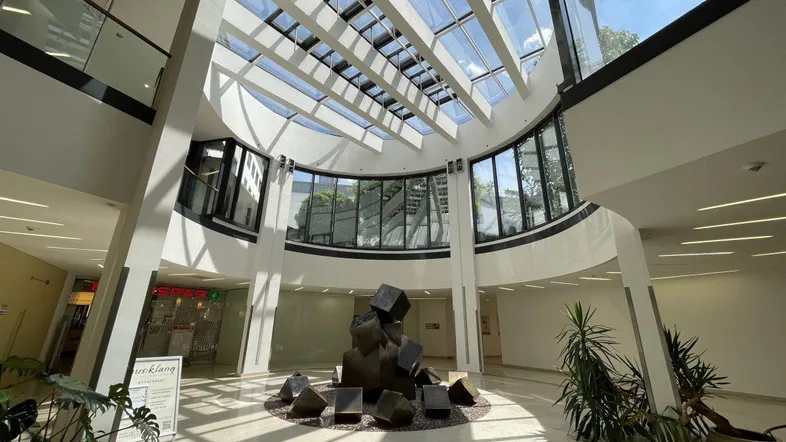 Helles, modernes Atrium mit Glasdach, das viel Tageslicht hereinlässt und eine Skulptur im Zentrum zeigt.