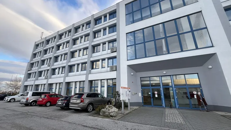 Modernes Bürogebäude mit mehreren Etagen und großen Fenstern, davor ein Parkplatz mit Autos.