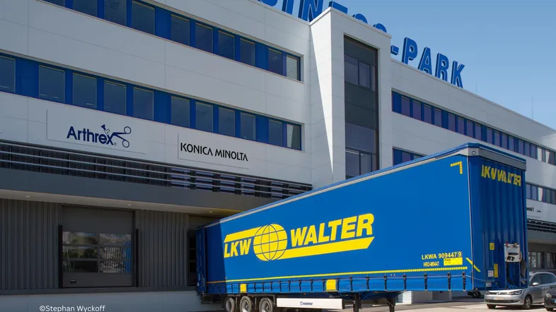Modernes Geschäftsgebäude mit Laderampe und einem LKW, der Waren entlädt. Die Fassade zeigt Firmenlogos.