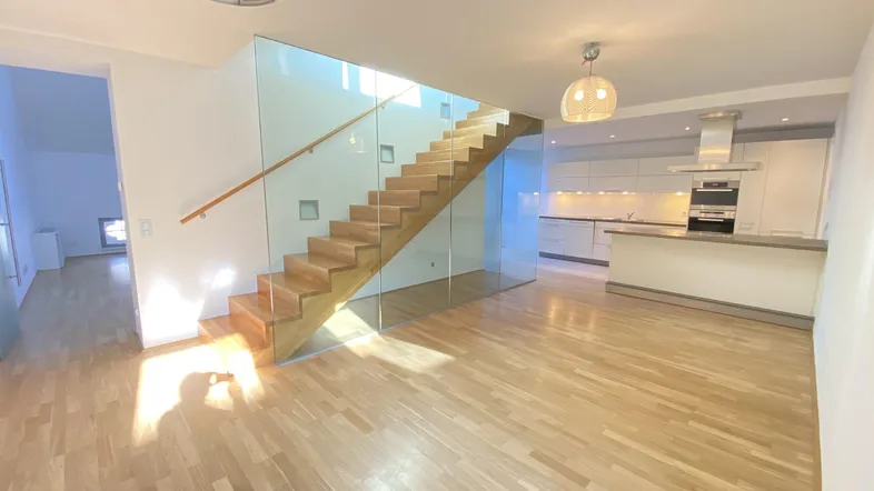 Helles, offenes Wohnzimmer mit Holztreppe, Glasgeländer und integrierter Küche im Hintergrund.