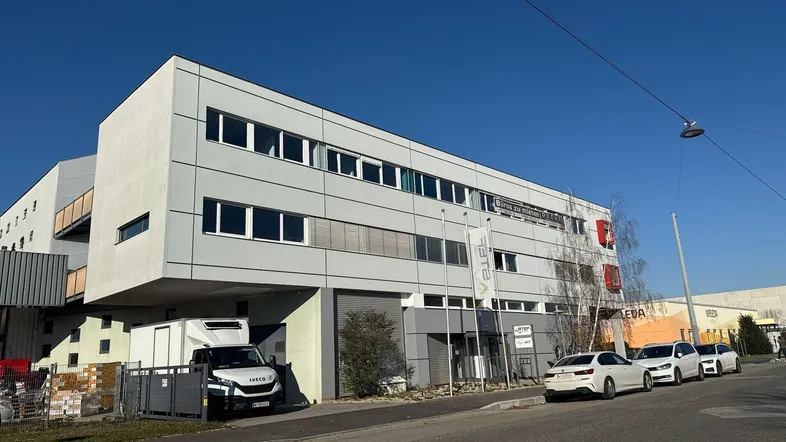 Modernes Bürogebäude mit mehreren Etagen, großen Fenstern und Parkplätzen davor.
