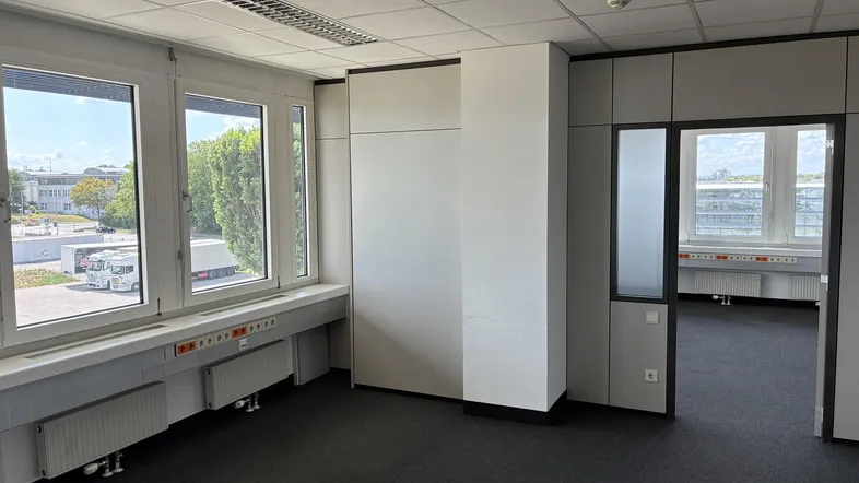 Geräumiges Büro mit Teppichboden, modularen Wänden und großen Fenstern mit Blick auf ein Gewerbegebiet.