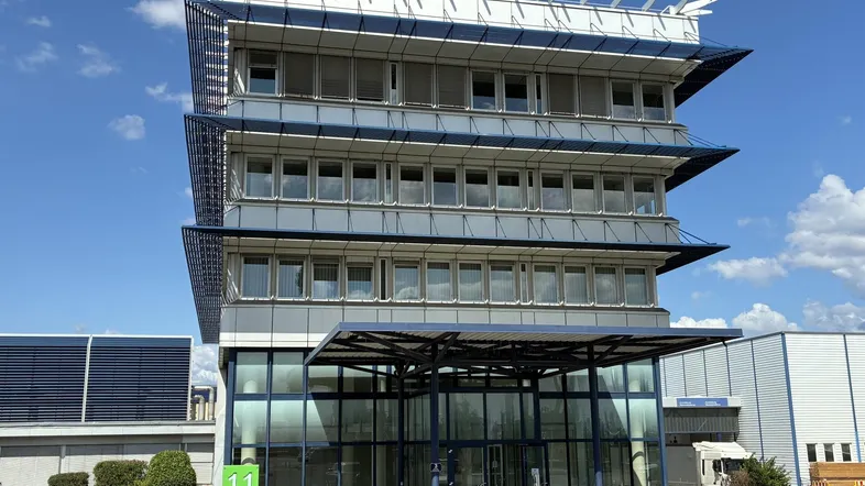 Modernes Bürogebäude mit mehreren Etagen, großen Fenstern und einer markanten Fassadenstruktur unter blauem Himmel.