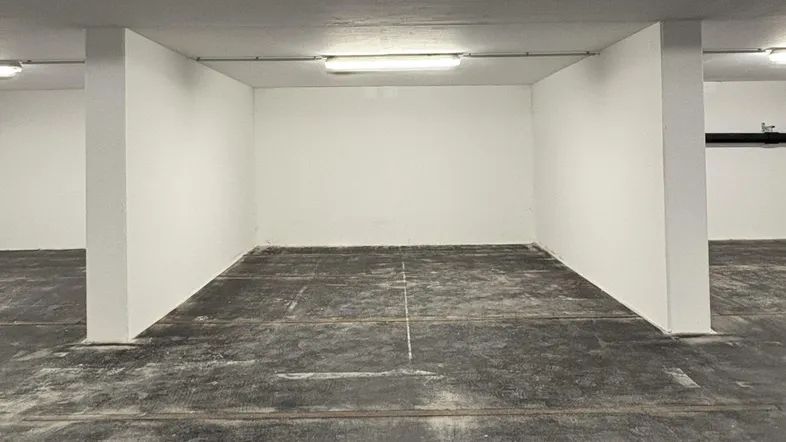 Leerer Tiefgaragenstellplatz mit Betonboden und weißen Wänden, beleuchtet von Neonröhren.