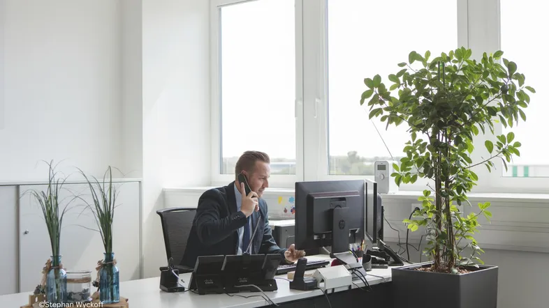 Ein Mann telefoniert in einem hellen Büro mit großen Fenstern und Pflanzen auf dem Schreibtisch.