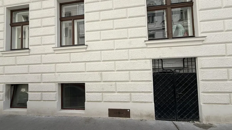 Fassade eines Altbaus mit weißem Putz, braunen Fenstern und einem Eingang zum Untergeschoss.
