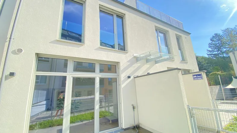 Moderne Gebäudefassade mit großen Fenstern, einer gefliesten Terrasse und einem kleinen Gartenbereich.