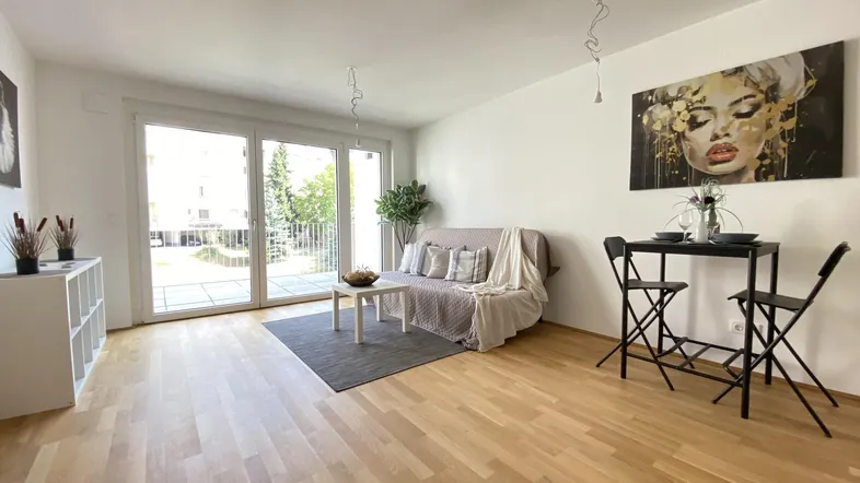 Helles Wohnzimmer mit Holzboden, Sofa und kleinem Essbereich, mit Zugang zu einem Balkon.