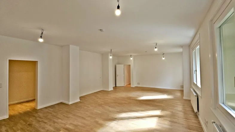 Geräumiger, leerer Raum mit hellem Holzfußboden, weißen Wänden und mehreren Fenstern für viel Tageslicht.
