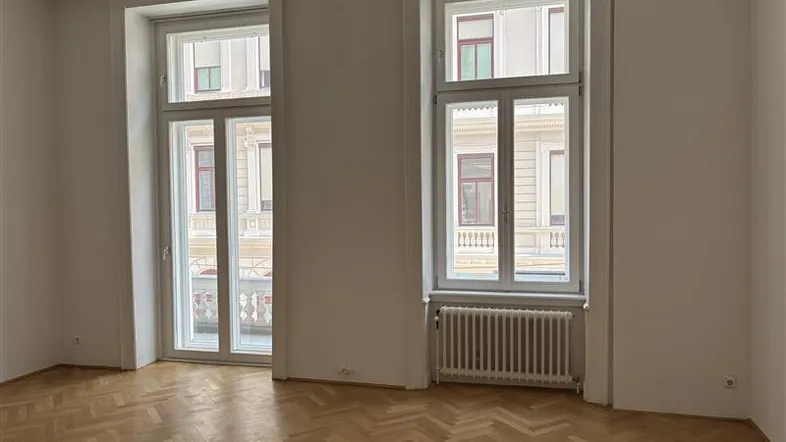 Heller Raum mit Fischgrätparkett, hohen Fenstern und Zugang zu einem Balkon, ideal für Wohn- oder Schlafzimmer.