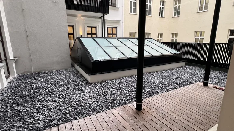 Außenbereich mit Holzterrasse, Kiesfläche und großem Dachfenster, umgeben von Gebäudefassaden.