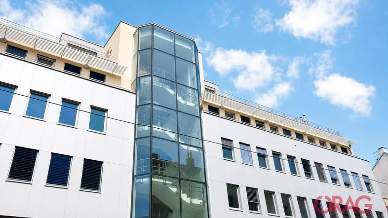 Moderne Gebäudefassade mit einer auffälligen Glasfassade und einem externen Aufzugsschacht unter blauem Himmel.