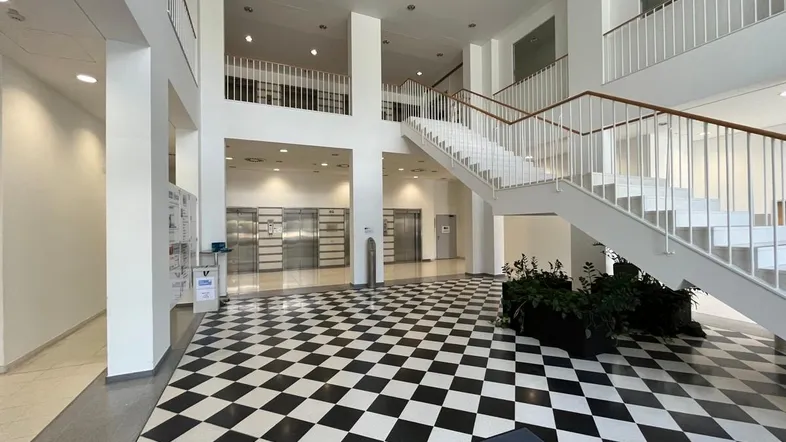 Geräumige Lobby mit markantem Schachbrettmuster-Boden, breiter Treppe und mehreren Aufzügen für den Zugang zu den Etagen.