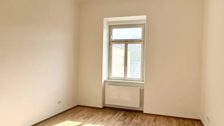 Helles, leeres Zimmer mit Parkettboden und einem Fenster, das viel Tageslicht hereinlässt.