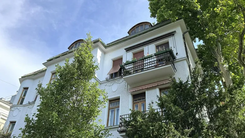 Klassisches Altbaugebäude mit weißer Fassade, Balkonen und grünen Bäumen unter einem strahlend blauen Himmel.