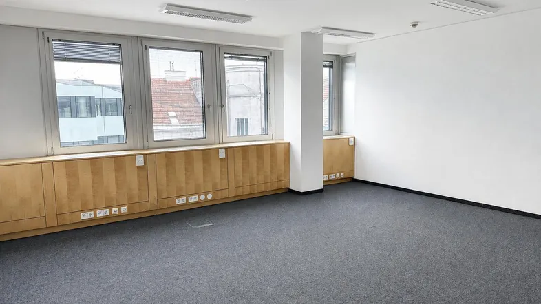 Geräumiges, leeres Büro mit Teppichboden, großen Fenstern und integrierten Holzschränken entlang der Wand.