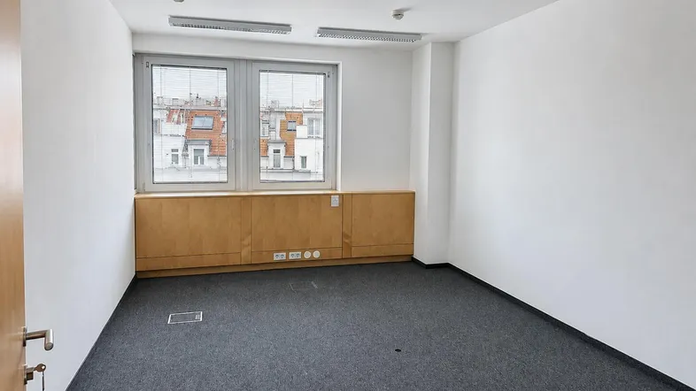 Heller Büroraum mit Teppichboden, großen Fenstern und integrierten Holzschränken unter der Fensterbank.