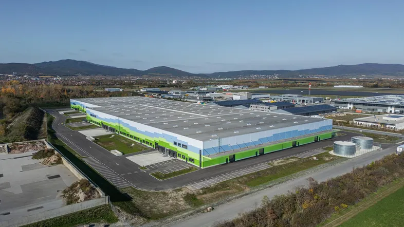 Moderne Industriehalle mit farbiger Fassade und weitläufigen Parkplätzen, umgeben von ländlicher Landschaft und Bergen.