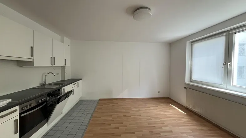 Heller Wohn- und Kochbereich mit integrierter Küchenzeile, Laminatboden und Fenster mit Blick nach draußen.