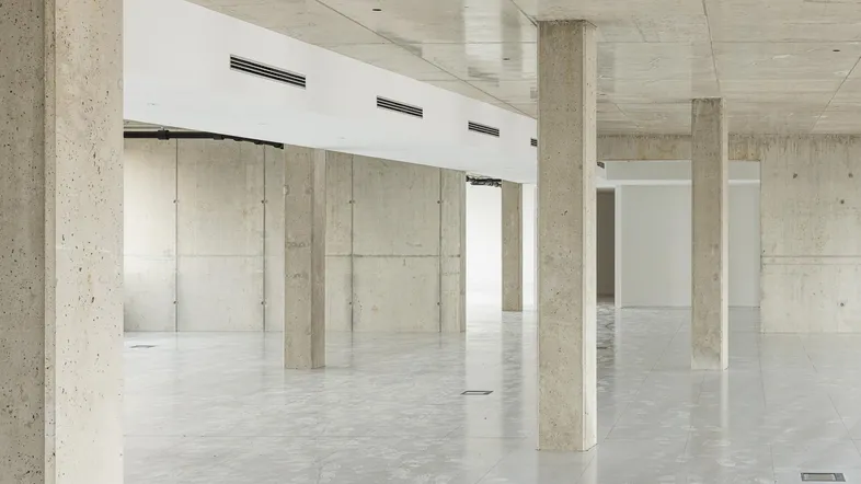 Großer, leerer Raum mit Betonpfeilern und -decke, hellgrauen Fliesen und sichtbaren Lüftungsschlitzen.
