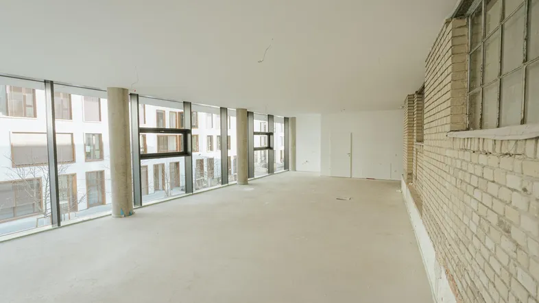 Großer, leerer Raum mit Betonpfeilern, einer Ziegelwand und einer Fensterfront zum Innenhof.