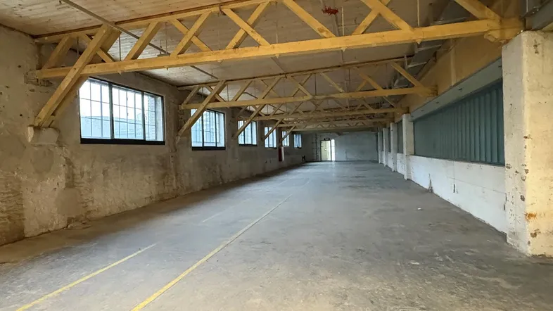 Großer, leerer Industrieraum mit Betonboden, sichtbaren Holzbindern und hohen Decken, gut beleuchtet.