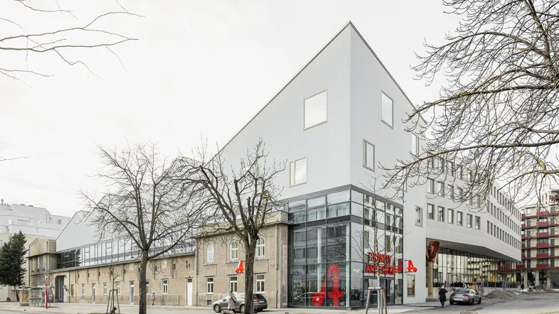 Moderne Architektur mit weißer Fassade und großen Glasflächen, daneben ein älteres Backsteingebäude an einer Straße.