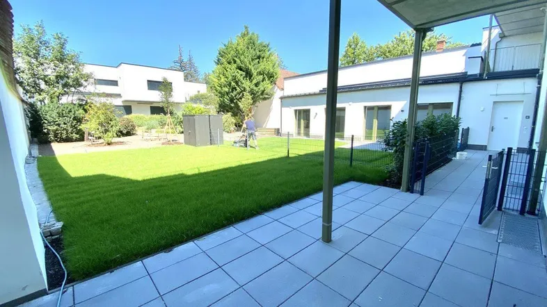 Große gepflasterte Terrasse mit Überdachung und angrenzendem grünen Rasenbereich in einer modernen Anlage.