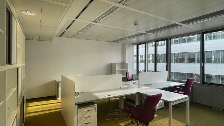 Helles Büro mit mehreren Arbeitsplätzen, Teppichboden und großen Fenstern mit Blick auf umliegende Gebäude.