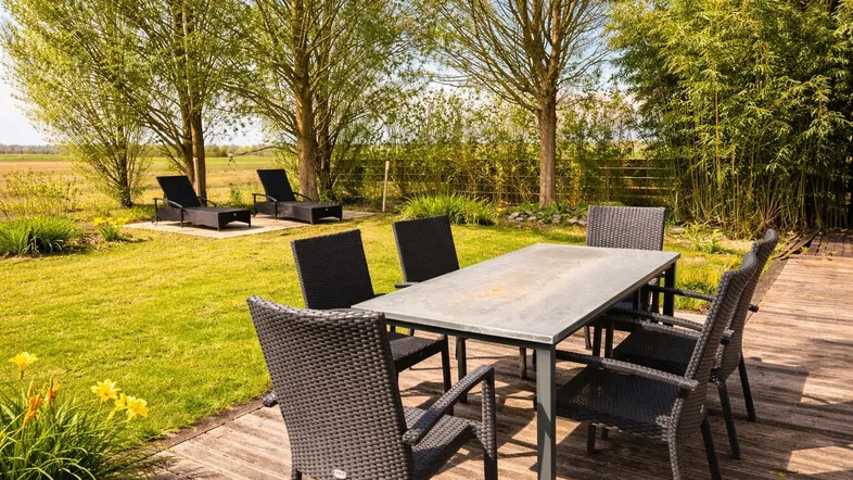 Große Holzterrasse mit Esstisch und Sonnenliegen, umgeben von Grünflächen und Bäumen.