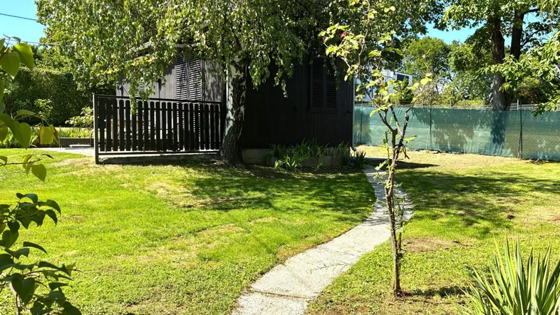 Grüner Garten mit einem kleinen Pfad, der zu einem dunklen Gartenhaus unter einem Baum führt.