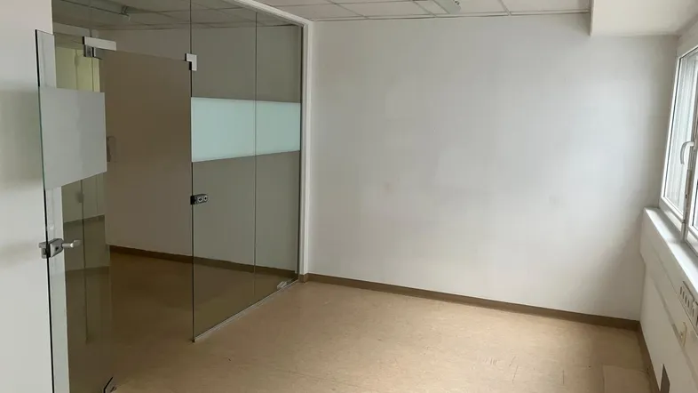 Leeres Bürozimmer mit modernen Glaswänden, hellen Böden und einem Fenster, das Tageslicht hereinlässt.