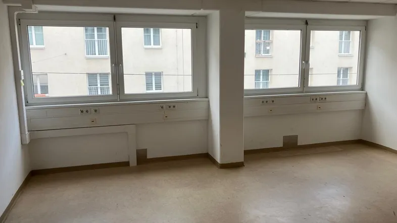 Heller Büroraum mit mehreren Fenstern, Blick auf gegenüberliegende Gebäude und abgehängter Decke.