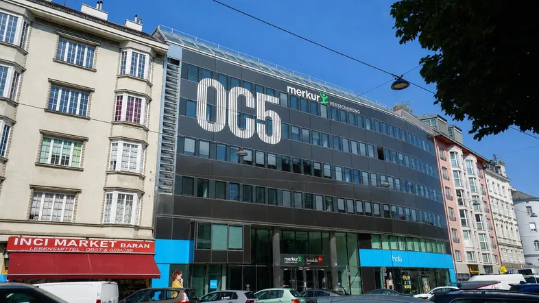 Modernes Bürogebäude mit Glasfassade und Beschriftung in einer städtischen Umgebung unter blauem Himmel.
