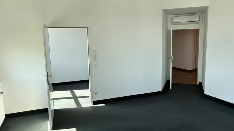 Heller Büroraum mit dunklem Teppichboden, weißen Wänden und zwei offenen Türen zu weiteren Räumen.