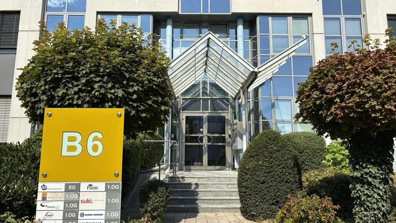 Modernes Bürogebäude mit Glasfassade und gepflegtem Eingangsbereich, umgeben von Grünflächen und einem Firmenschild.