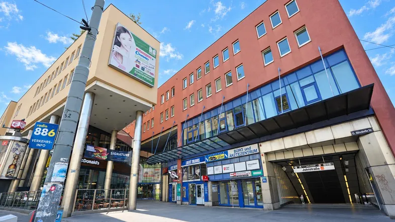 Moderne Geschäftsgebäude mit U-Bahn-Zugang und verschiedenen Ladenlokalen in einer belebten Stadtumgebung.