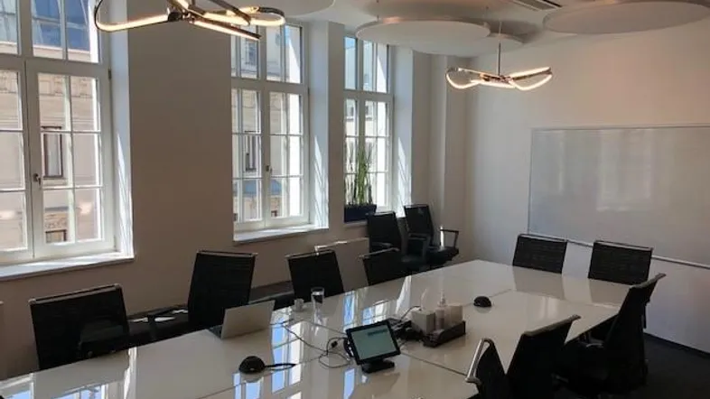 Heller Konferenzraum mit großem weißen Tisch, modernen Deckenleuchten und hohen Fenstern, die viel Tageslicht hereinlassen.