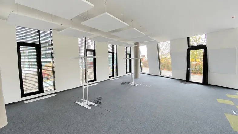 Großer, heller Büroraum mit grauem Teppichboden, vielen Fenstern und schwebenden Akustikpaneelen.